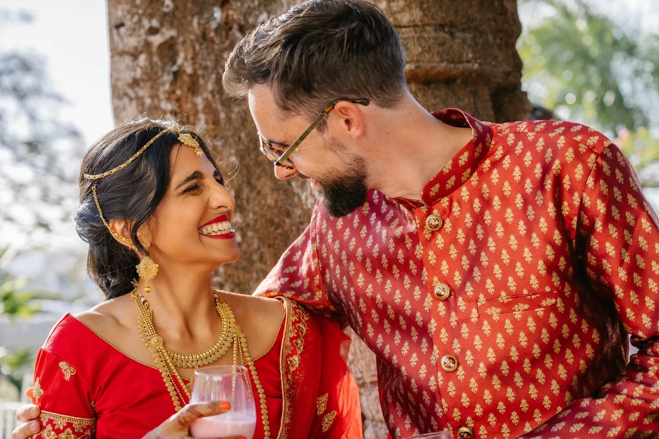 Les mariés en tenue indienne rouge se regardent en riant, un verre à la main