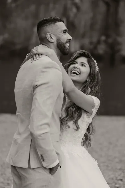En noir et blanc, la mariée rit aux éclats dans les bras du marié en costume clair