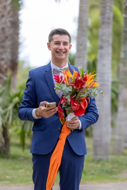 Le marié en costume bleu sourit, un bouquet de fleurs tropicales aux rubans orange dans les mains
