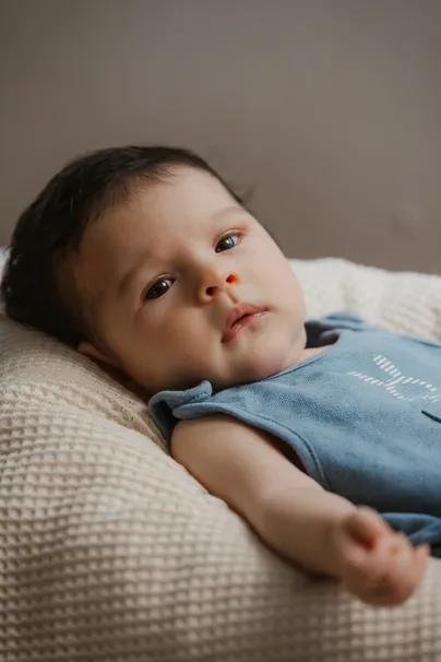 Le bébé en barboteuse bleue est allongé sur un coussin tricoté, le regard vers l’objectif