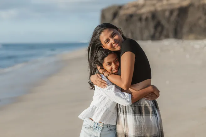 Une mère serre sa fille contre elle en souriant, sur une plage au pied d’une falaise