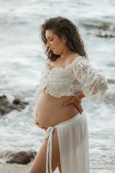 Future maman en haut de dentelle et jupe blanche, tête baissée vers son ventre, face à la mer