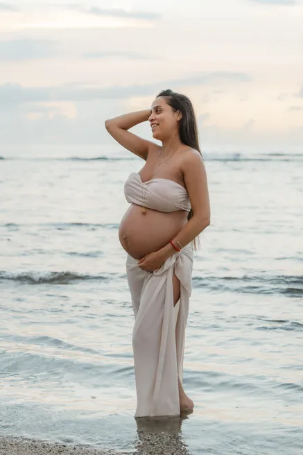 Future maman drapée de beige debout dans l’eau, main dans les cheveux, regard vers le large