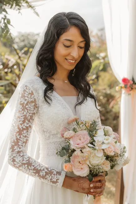 La mariée baisse les yeux sur son bouquet de roses pâles, manches de dentelle