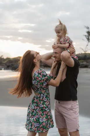 Une petite fille rit sur les épaules de son père, que sa mère embrasse sur la plage