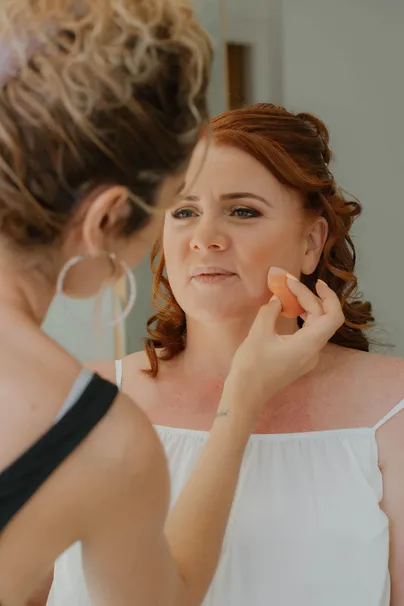 La mariée aux cheveux roux se fait maquiller à l’éponge, le regard au loin