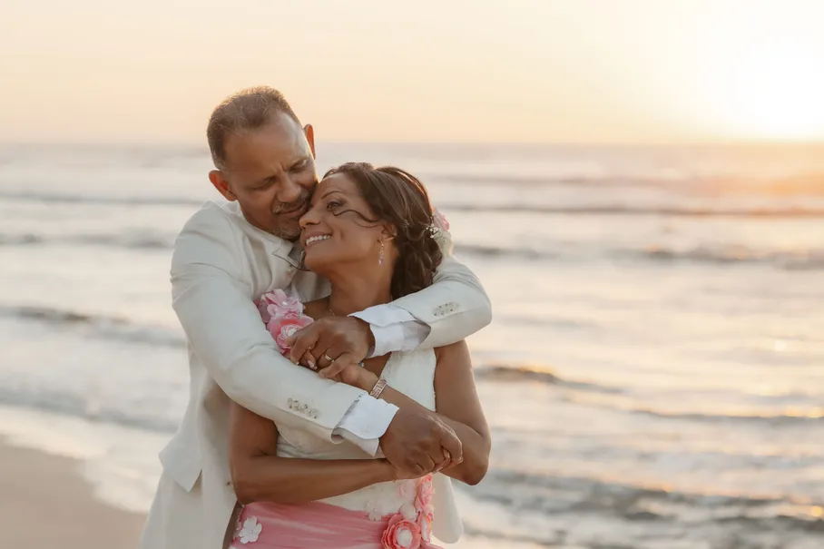 Un couple enlacé rit face à la mer au coucher du soleil, lui en costume clair, elle en robe rose