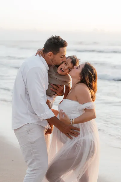 Le papa porte leur enfant et embrasse la future maman en robe blanche, face à la mer