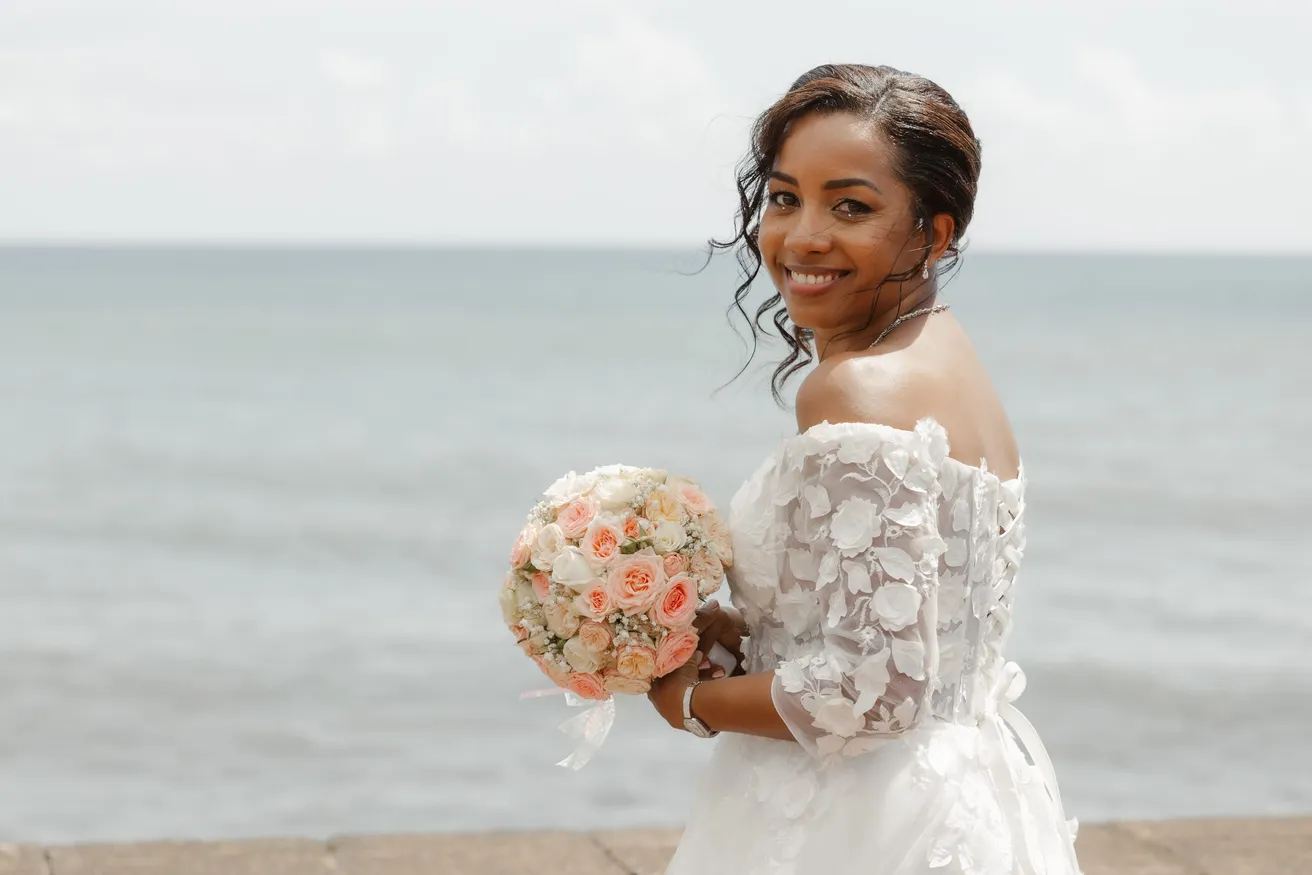 La mariée sourit par-dessus l’épaule face à la mer, bouquet pastel et manches de dentelle fleurie