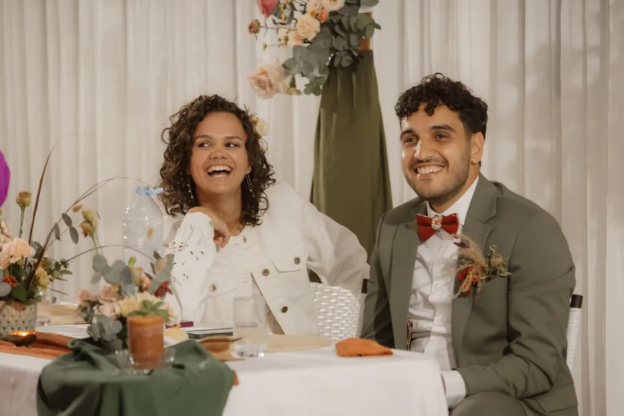 Les mariés rient, assis à leur table de réception décorée de fleurs et de lin vert