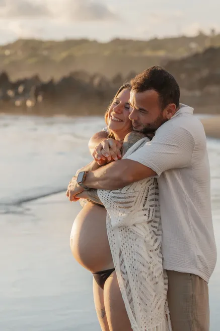 Un couple enlacé en riant au bord de l’eau, la future maman en tunique blanche ajourée