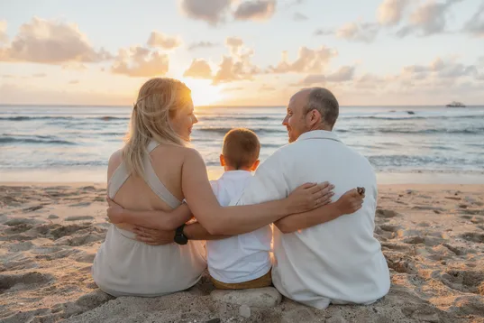 Un couple et leur fils assis de dos sur un rocher, face au soleil couchant