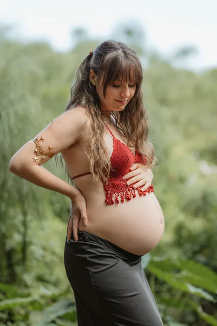 Future maman de profil, haut rouge crocheté et ventre nu, devant la végétation
