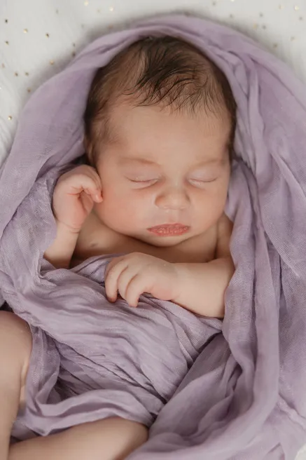 Le nouveau-né dort emmailloté dans un voile mauve, les mains repliées sous le menton
