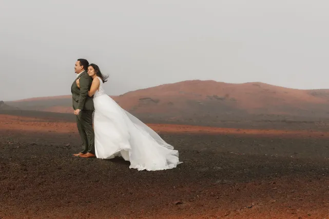 Blottie dans le dos du marié, la mariée pose dans le vent, sa robe soulevée, devant un cône volcanique rouge