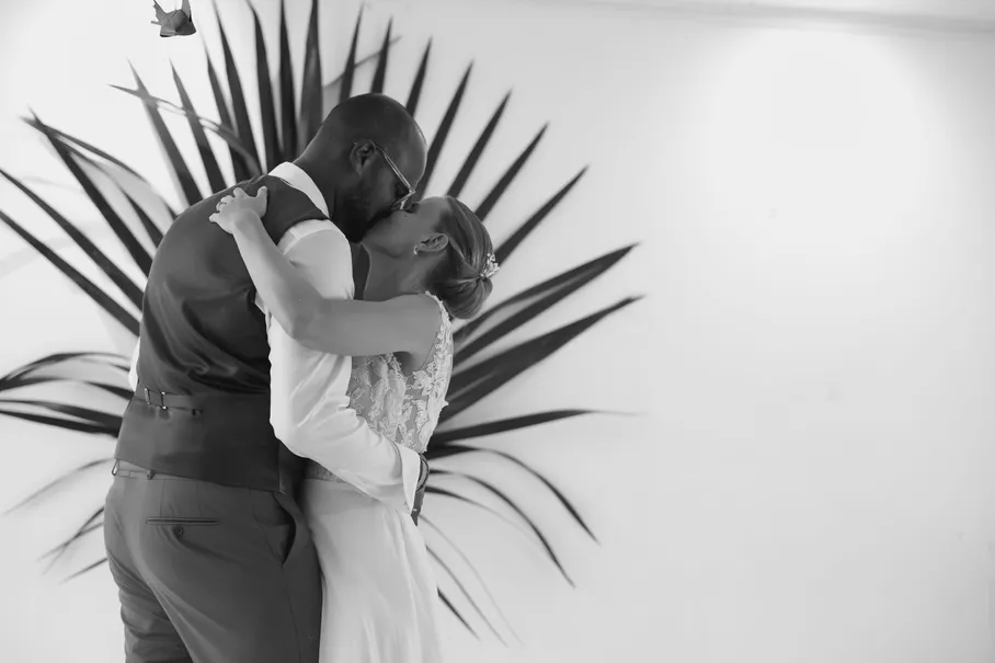 En noir et blanc, les mariés s’embrassent devant une grande palme en éventail