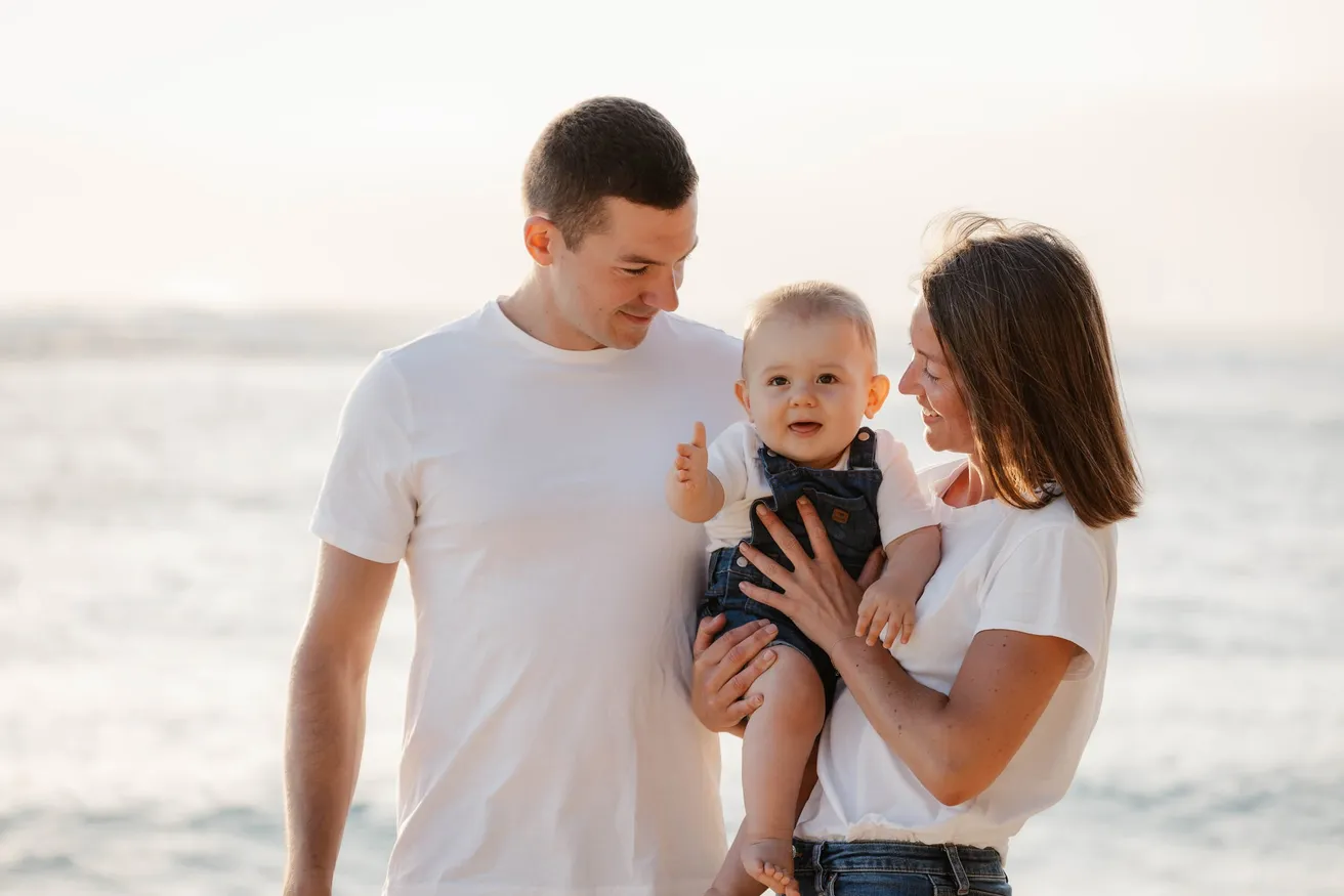 Un couple en tee-shirts blancs entoure son bébé au bord de la mer, dans la lumière du soir