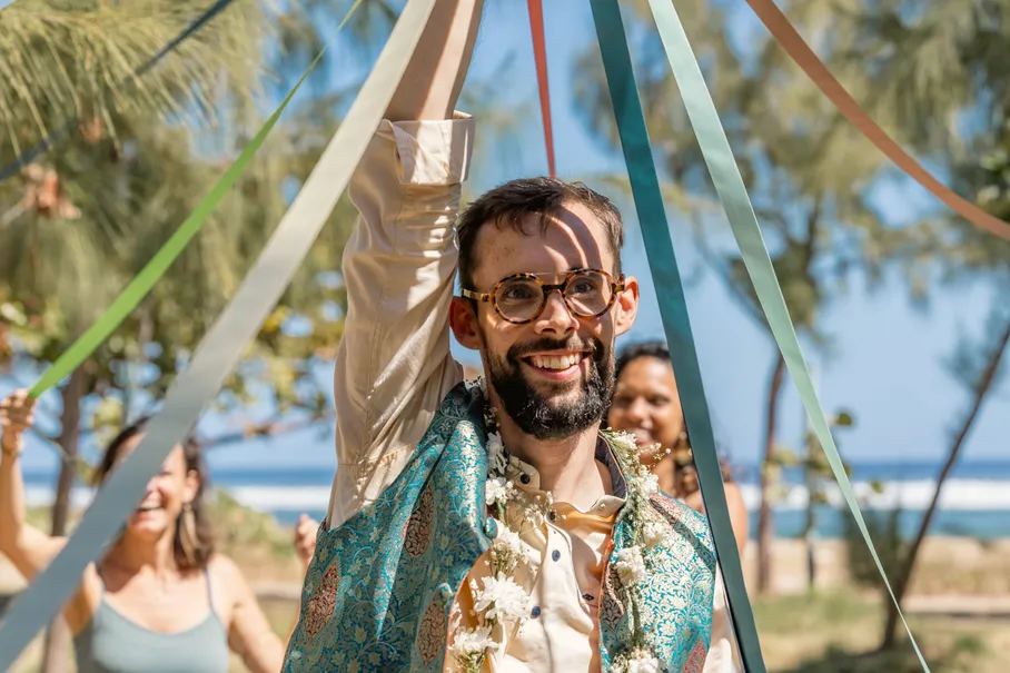 Le marié en veste turquoise brodée lève le bras sous les rubans colorés, collier de fleurs au cou