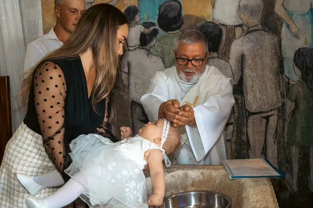 Un prêtre baptise une petite fille en robe blanche au-dessus d’une bassine, devant ses parents