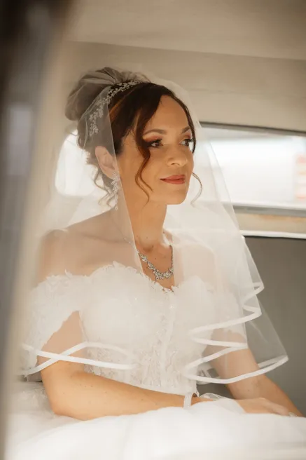 La mariée en voile et robe bustier, assise près d’une fenêtre, le regard au loin