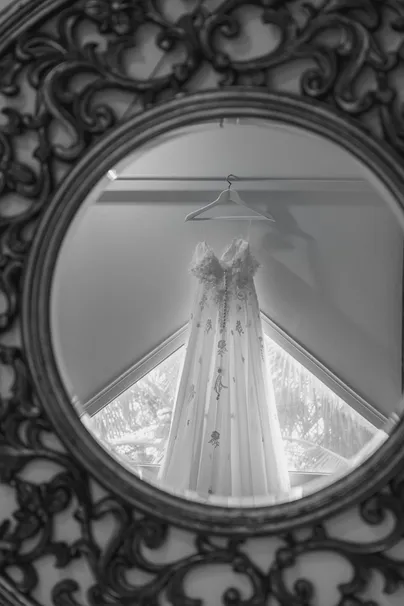 En noir et blanc, la robe de mariée suspendue, vue à travers un miroir rond en fer forgé