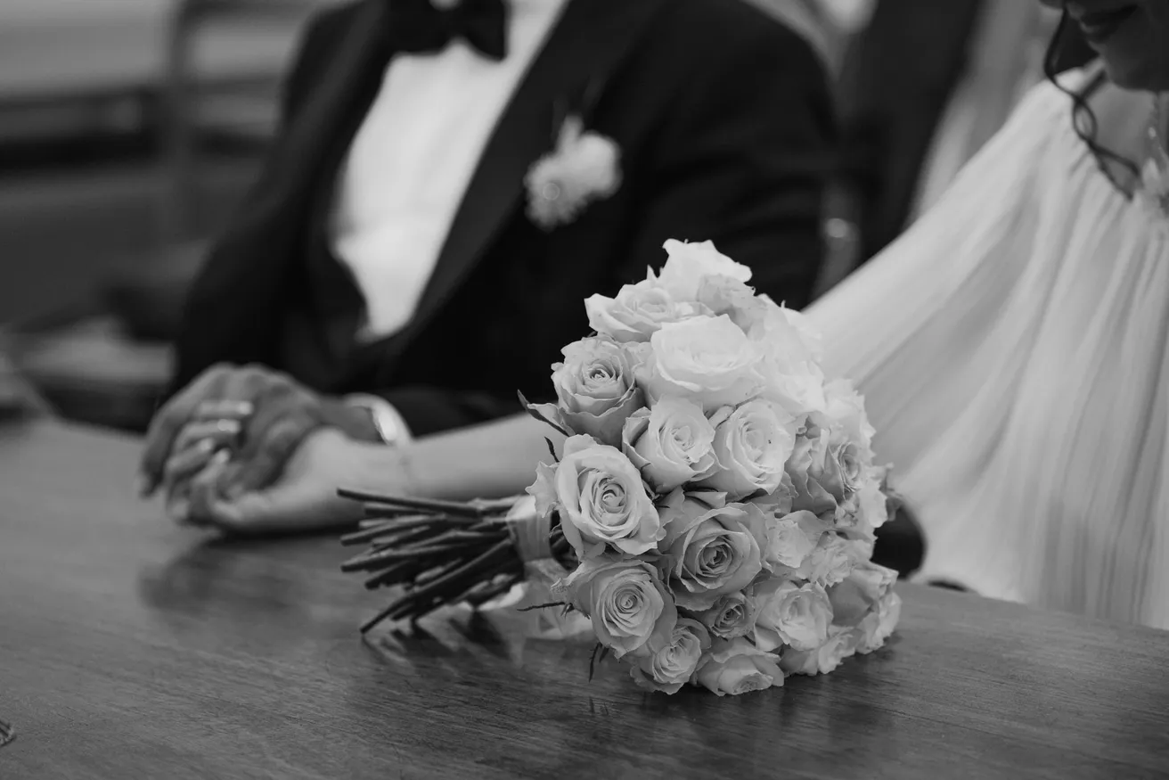 En noir et blanc, mains jointes des mariés sur une table, bouquet de roses posé à côté