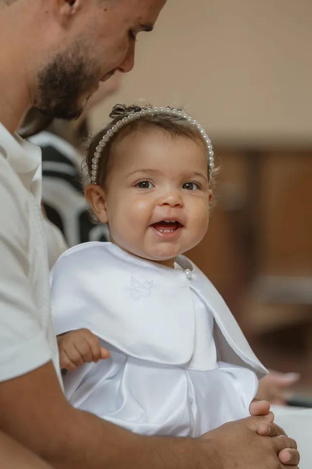 Un bébé en tenue blanche, un bandeau dans les cheveux, sourit dans les bras de son papa