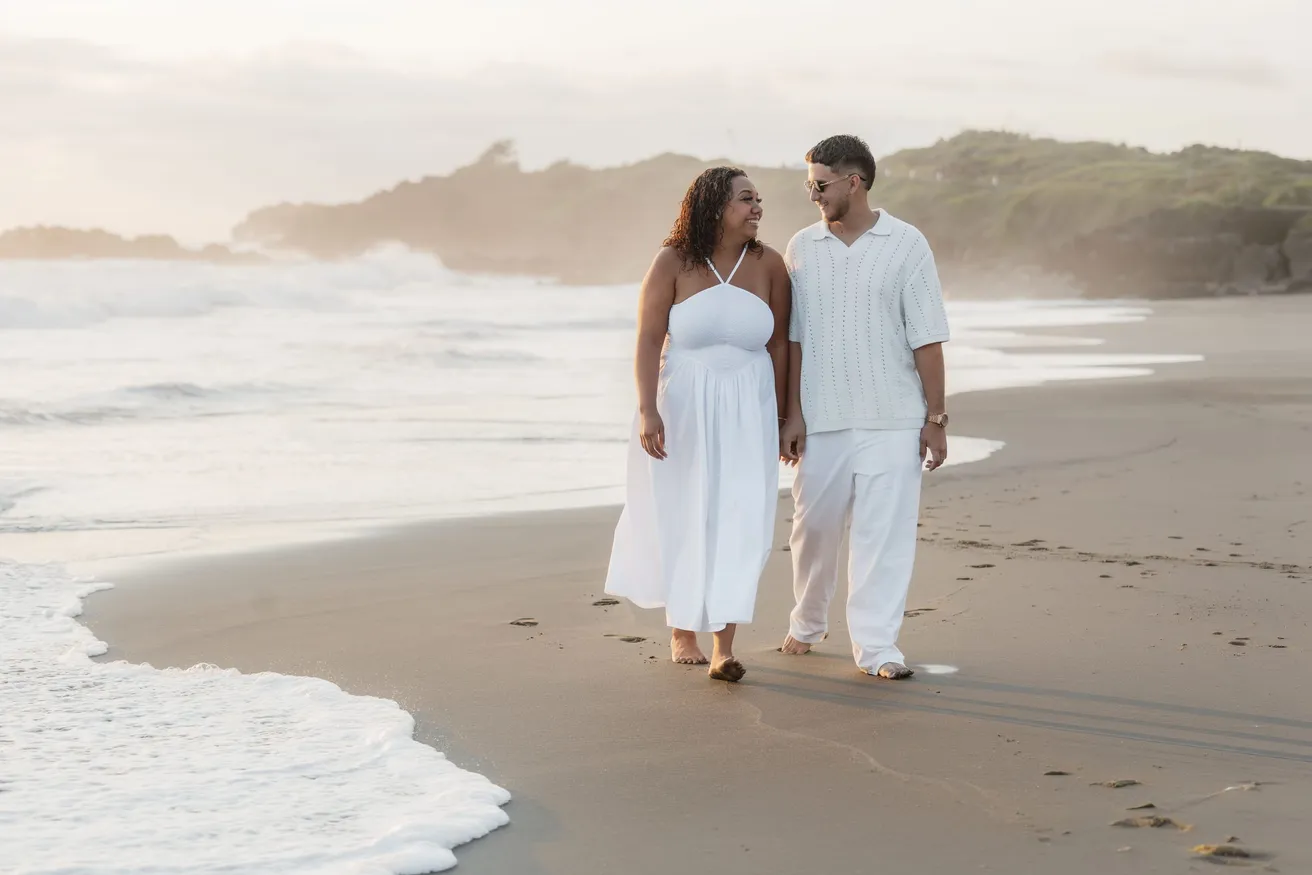 Un couple en blanc marche sur le sable sombre au bord de l’écume, collines verdoyantes au fond