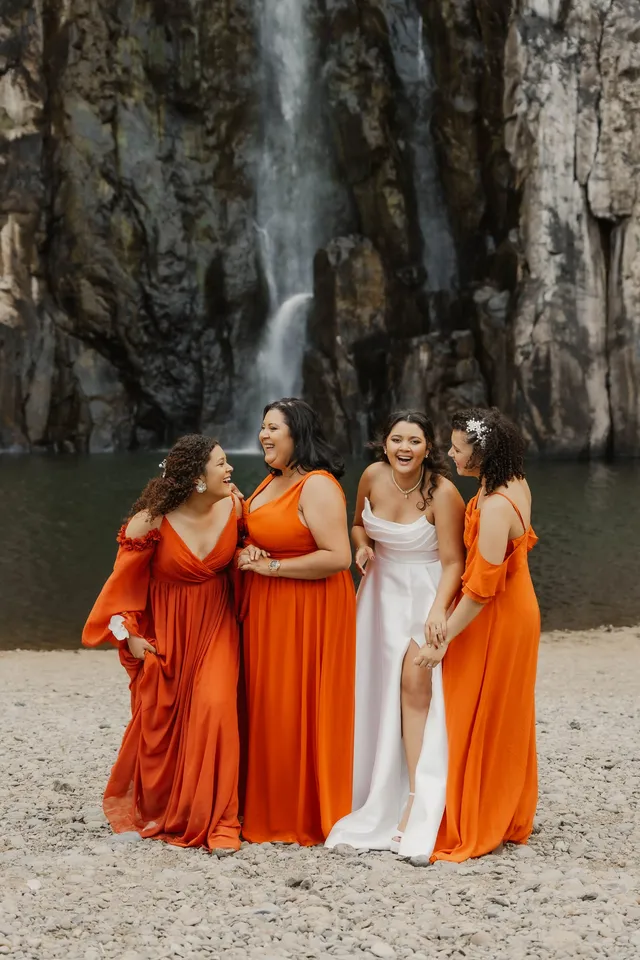 La mariée rit avec ses trois demoiselles d’honneur en robes orange, au pied d’une cascade
