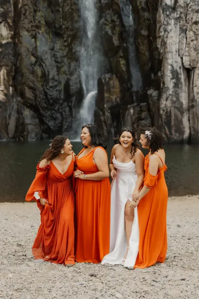 La mariée rit avec ses trois demoiselles d’honneur en robes orange, au pied d’une cascade