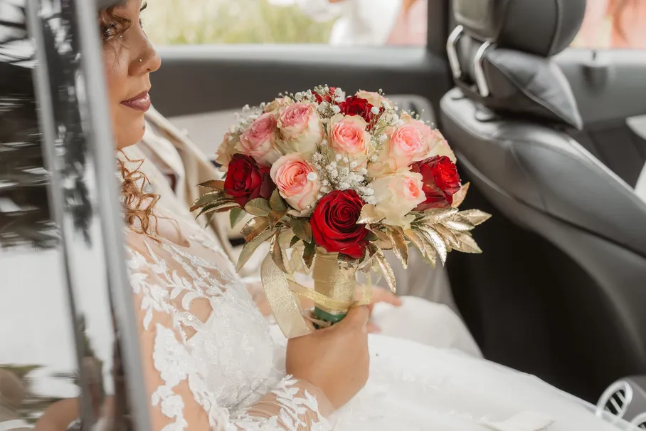 La mariée assise en voiture, bouquet de roses rouges et roses aux feuilles dorées sur les genoux