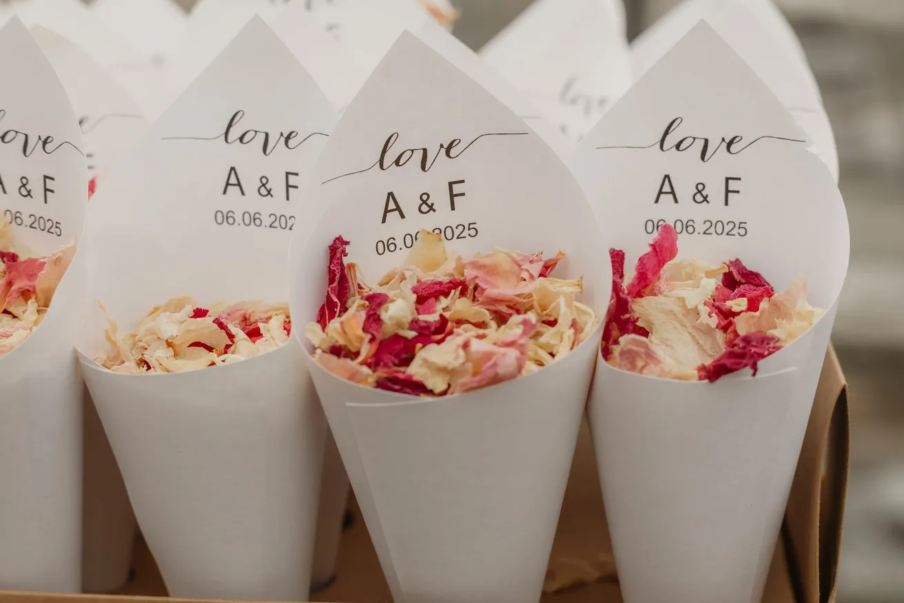 Cornets de papier blanc remplis de pétales de roses séchés, alignés pour la sortie des mariés