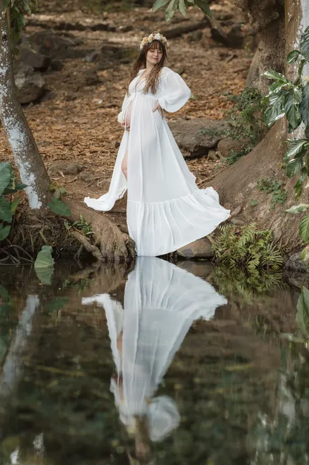 Future maman en robe blanche au bord d’un bassin en sous-bois, reflétée dans l’eau
