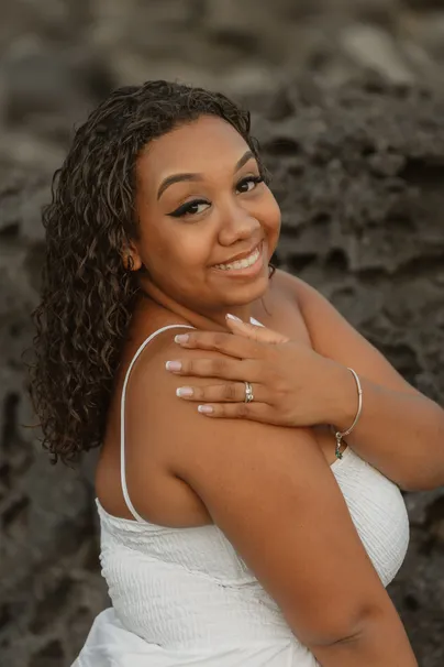 Une jeune femme aux cheveux bouclés sourit, la main sur l’épaule, devant une roche volcanique