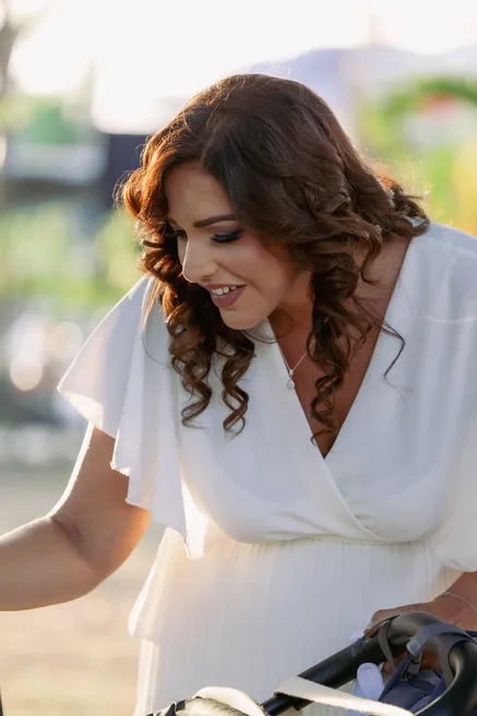 Invitée en robe blanche penchée en souriant au-dessus d’une poussette, dans la lumière du soir