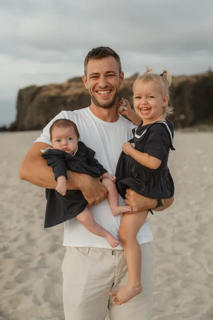 Un père rit en portant ses deux petites filles vêtues de noir, sur le sable devant la falaise