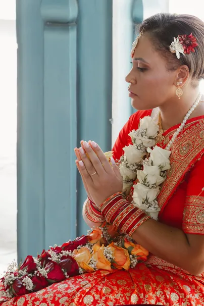 La mariée en sari rouge, mains jointes en prière, une guirlande de fleurs blanches au cou