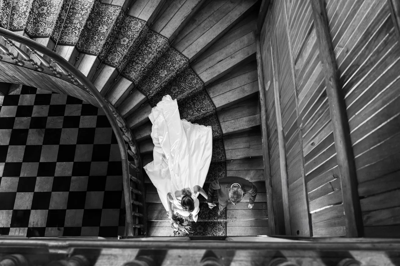 En noir et blanc, vue plongeante sur un escalier en colimaçon où descend la mariée en voile