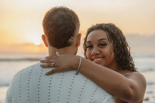 Au coucher du soleil, elle enlace son fiancé face à la mer et sourit, la bague au doigt