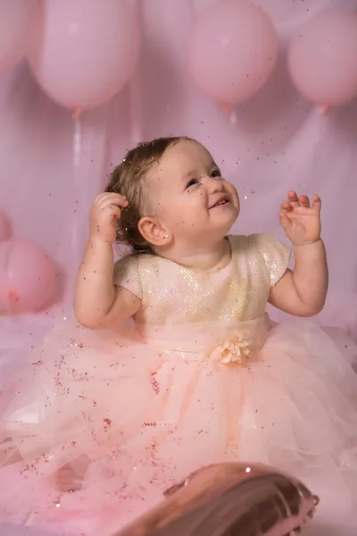 Une petite fille en robe de tulle rose lève les mains en riant, sous les ballons et les confettis