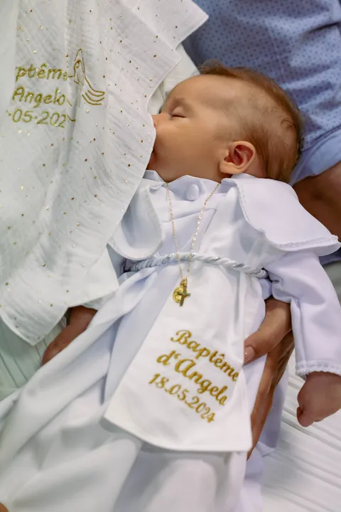 Un bébé endormi en tenue blanche, son étole de baptême brodée de fil doré posée sur lui